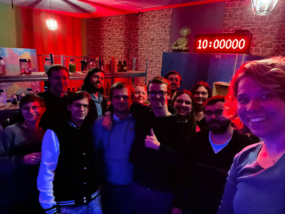 Zu sehen ist ein Gruppenfoto mit 12 Personen. Alle posieren für die Kamera. Im Hintergrund ist ein mittelgroßer Raum zu sehen mit dekorativen Labortutensilien. Der ganze Raum ist rot beleuchtet.