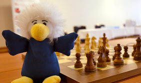 Das Maskottchen Chessy sitzt vor einem aufgebauten Schachbrett