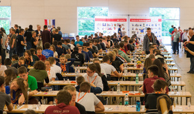 Foto von dem Spielsaal der Meisterschaften 2019. Zu sehen sind unzählige Tischreihen bis zum Hintergrund, die von links nach rechts verlaufen. In jeder Reihe sitzen sich 6 Jugendliche an Schachbrettern gegenüber. 