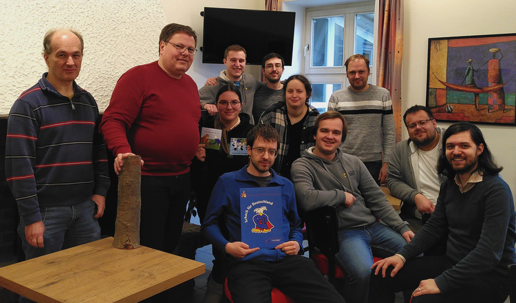 Gruppenfoto der Klausurtagung Gruppenfoto im Seminarraum der Jugendherberge. Zu sehen sind die 11 Teilnehmenden, 4 Sitzen auf Stühlen in der vorderen Reihe und 7 Stehen dahinter. Alle schauen in die Kamera
